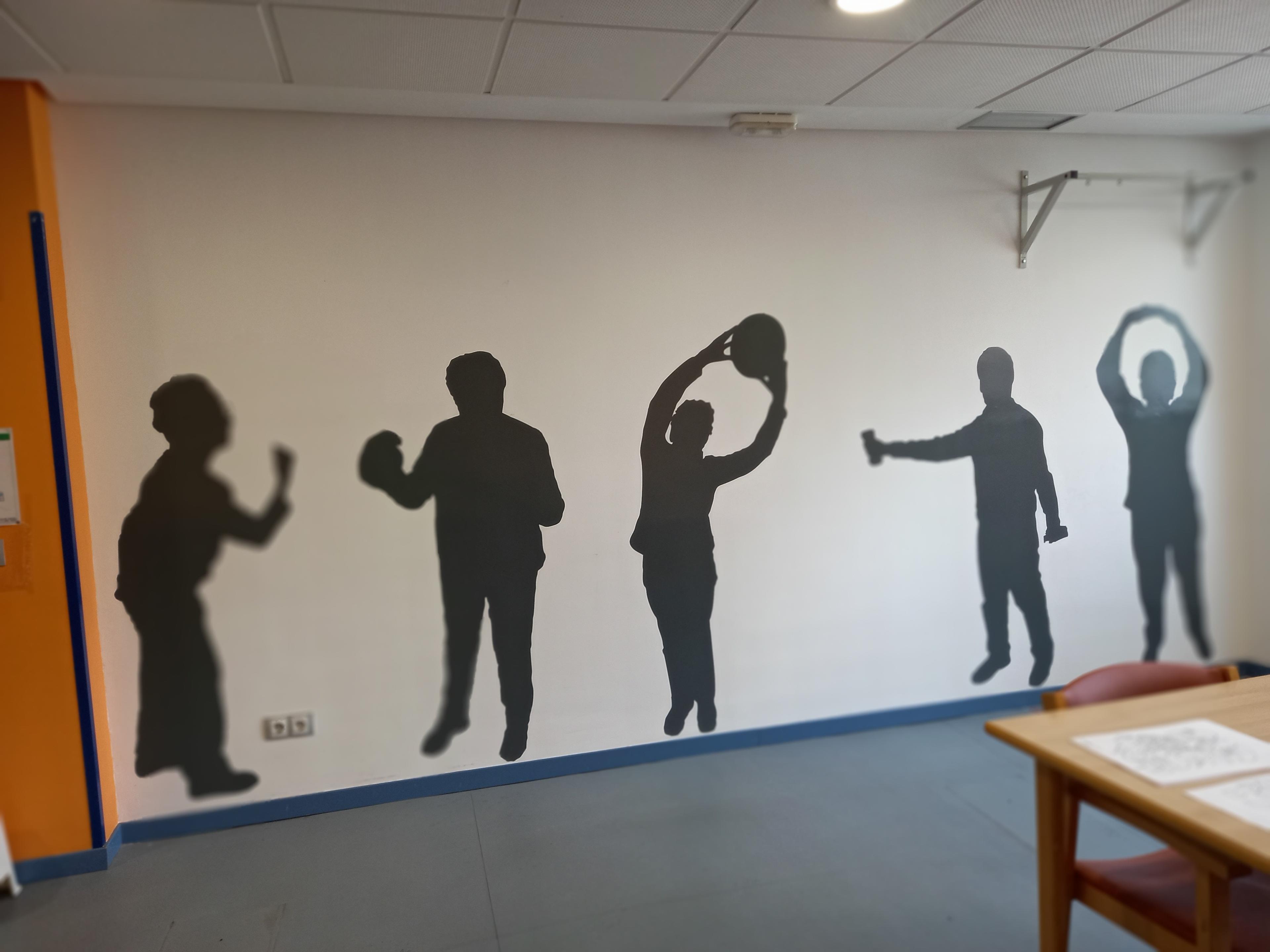 Mural en pared de centro de cuidados con siluetas de personas realizando diferentes ejercicios: estiramiento, levantamiento de pesas y movimientos de brazos. Diseño estimula actividad física y memoria motora.