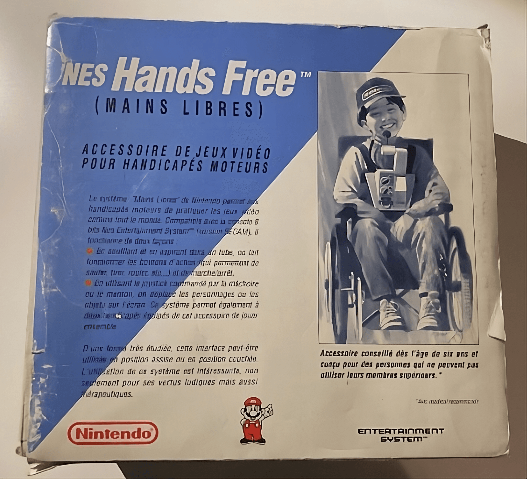 Caja del NES Hands Free, un accesorio de Nintendo diseñado para personas con discapacidades motoras. La imagen en la caja muestra a un niño en una silla de ruedas utilizando el dispositivo, que le permite jugar sin usar las manos