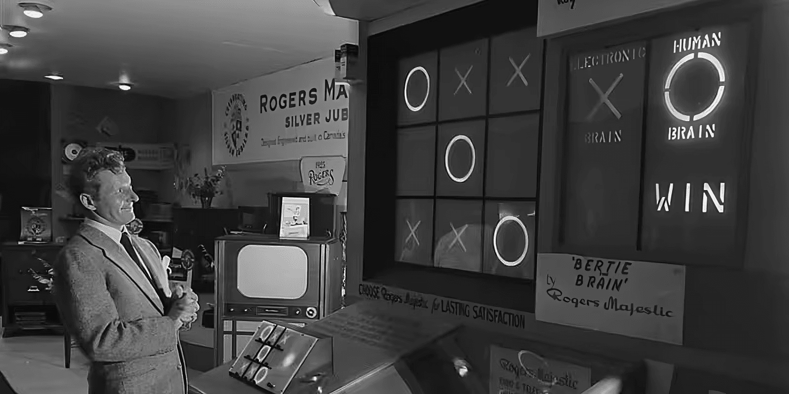 Un hombre de pie frente a 'Bertie the Brain', una máquina de tic-tac-toe (tres en raya) de gran tamaño de la década de 1950, que muestra un juego en curso en una pantalla vertical con botones para seleccionar las jugadas