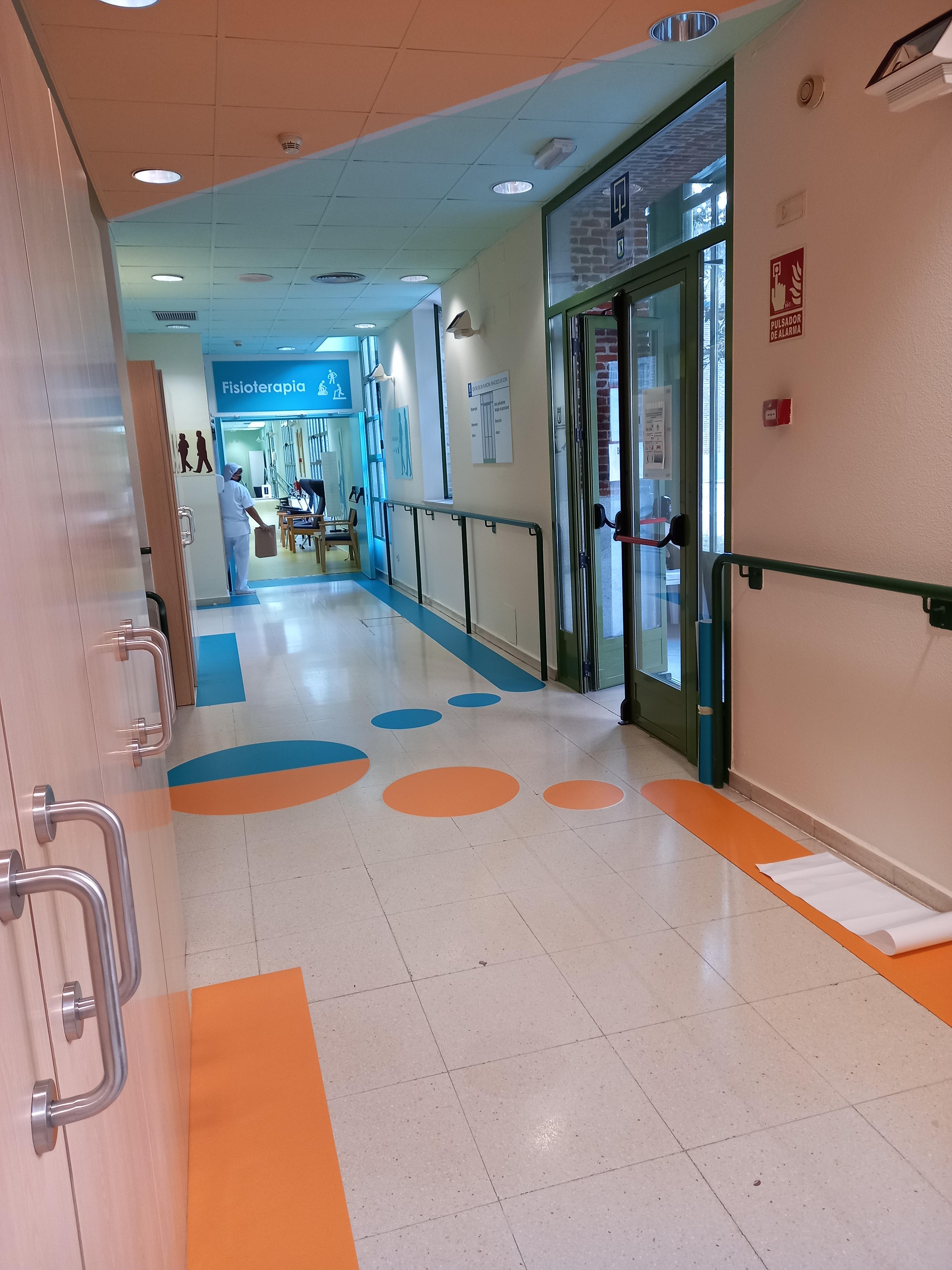 Pasillo de centro de salud con señalización en suelo: círculos azules y naranjas guían hacia fisioterapia. Barandillas y puertas amplias. Diseño facilita orientación y movilidad segura para mayores.