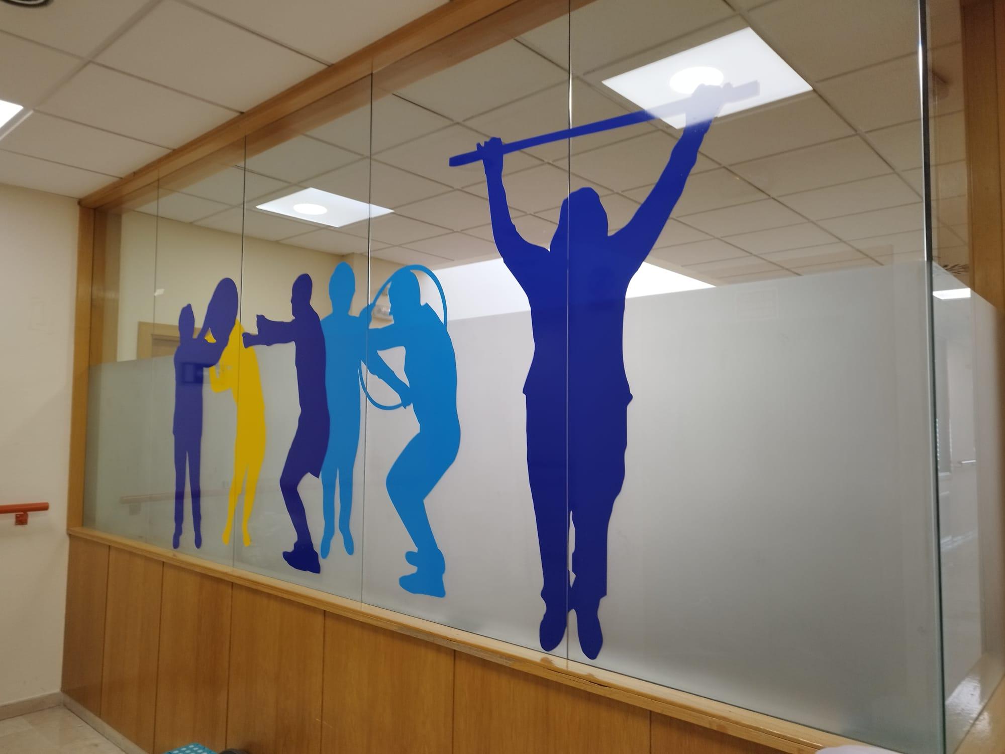 Panel de vidrio en centro de cuidados con siluetas coloridas de personas haciendo ejercicios. Figuras en azul y amarillo muestran estiramientos y movimientos. Diseño promueve actividad física y estimulación visual.