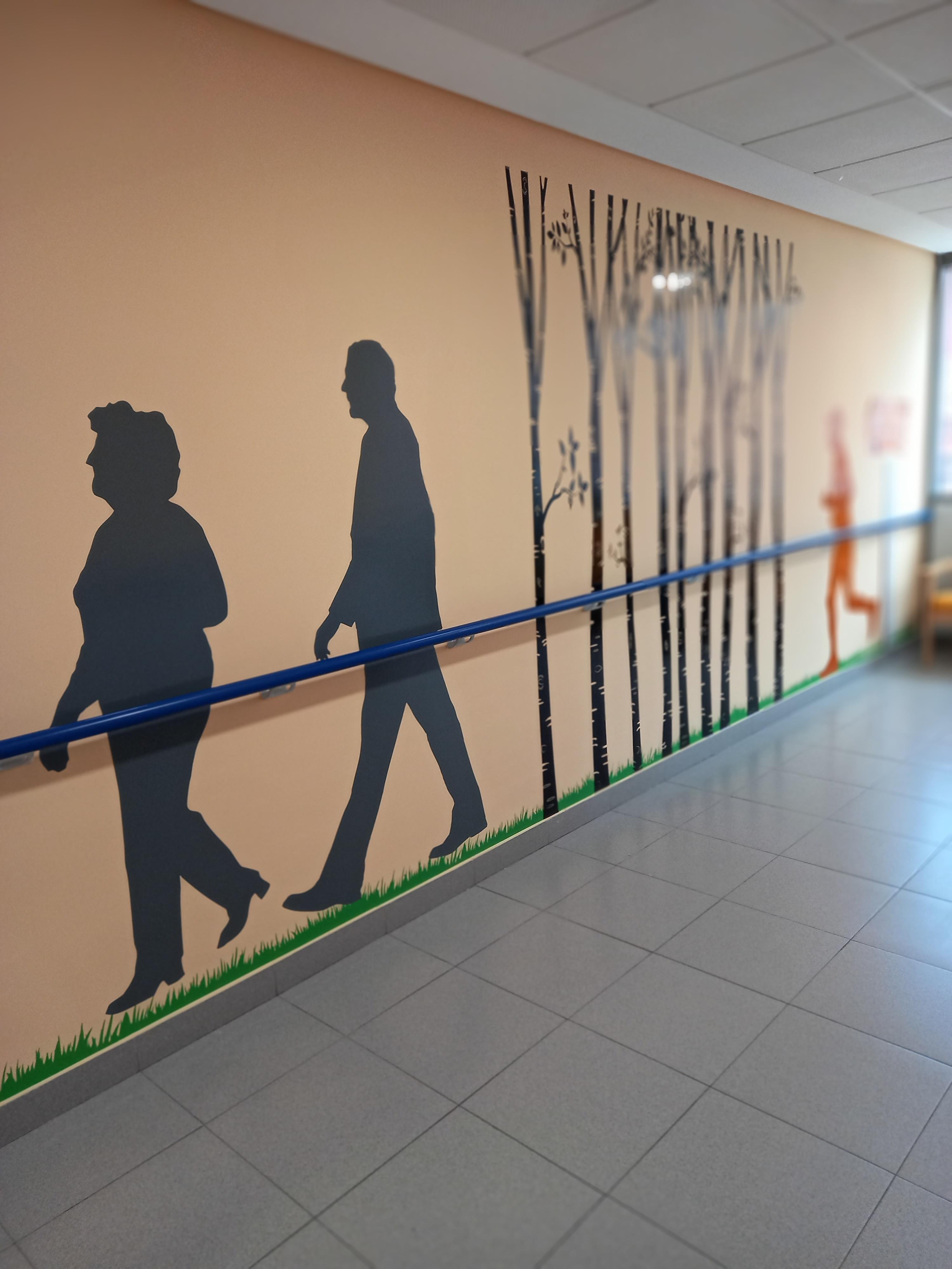 Pasillo con mural en la pared: siluetas de personas caminando, árboles y césped. Barandilla azul a lo largo del mural. Diseño estimula actividad y orientación espacial en centro de cuidados.